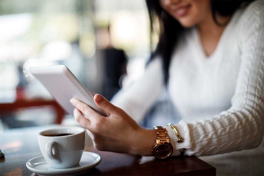Image of a woman’s hand holding a cell phone and a cup of coffee on the table.
